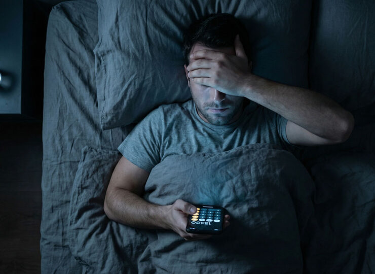Man lying awake in dark bedroom at night, face lit by phone screen showing calculator app, hand on forehead in frustration
