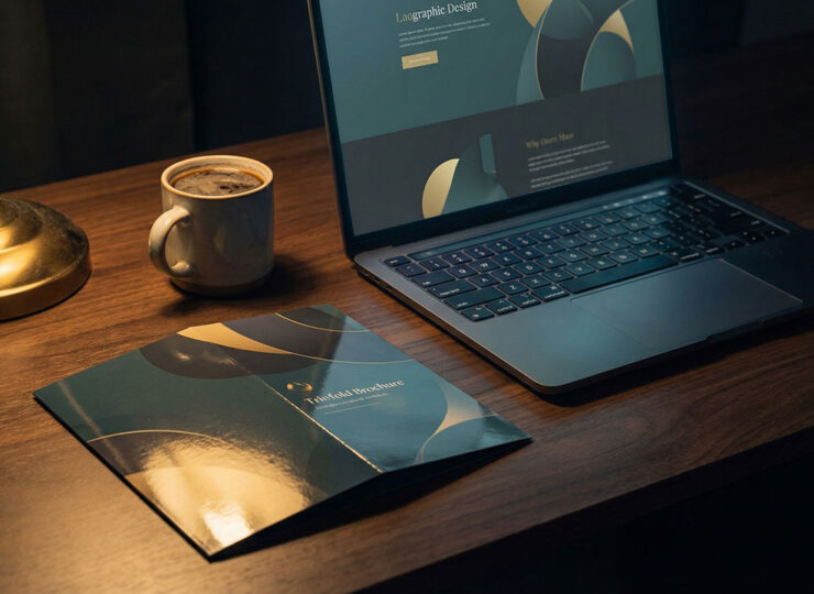 Open laptop showing a modern website beside an untouched glossy brochure on a dimly lit desk
