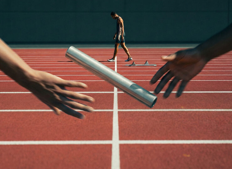 A relay baton suspended mid-air between two blurred hands during a failed handoff, with a solo runner in sharp focus already past the finish line in the background.